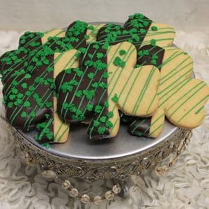 STP-001B St. Patrick's Day Sugar Cookies, shamrock-shaped and half dipped in chocolate with green sprinkles and lines, are arranged on a silver stand with bead accents on a white textured surface.