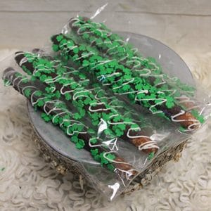 STP-028 Shamrock Pretzels: Chocolate-covered pretzel rods with green shamrock sprinkles and white drizzle, arranged on a silver plate, wrapped in clear plastic, and displayed on a decorative off-white textured surface.
