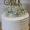 RC-003 is a white frosted cake adorned with white baby's breath flowers and a gold glitter "God Bless" topper with a cross, presented on a gold cake board.