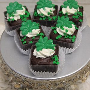 Six STP-025 Chocolate Bailey's Cup Gluten Free treats, topped with white frosting and green shamrock decorations, are displayed on a silver cake stand with a decorative edge.