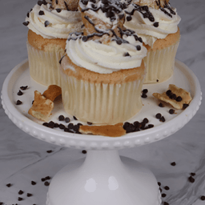 Three CC-000T Cannoli Cupcakes, topped with white frosting, mini chocolate chips, and cannoli pieces, sit on a white cake stand with chocolate chips and pastry crumbs scattered around.