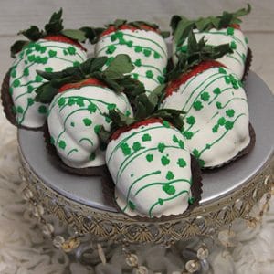 Six STP-021 White Chocolate dipped strawberries, decorated with green drizzle and clover-shaped sprinkles, are arranged on a silver beaded cake stand above white fabric petals.