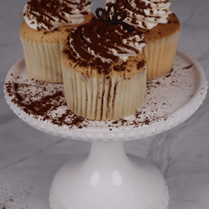 Three CC-000U Tiramisu Cupcakes dusted with cocoa and topped with chocolate decorations are displayed on a white, decorative cake stand against a light marble background.