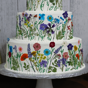 The WD-003 is a three-tiered white cake adorned with colorful, realistic edible flowers and green stems, showcased on a white cake stand against a light wooden background.