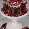 Three CC-000D Chocolate raspberry cupcakes sit on a white cake stand, topped with pink frosting, raspberries, and chocolate decorations. Chocolate shavings and raspberries are scattered around the stand.