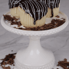 Three CC-000M Boston Cream cupcakes topped with chocolate glaze and white icing drizzle sit on a white cake stand, surrounded by chocolate shavings on the stand and marble surface.