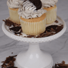 Three CC-000J Gold Oreo Cupcakes with cookies and cream frosting, each topped with a half-dipped chocolate cookie, are displayed on a white cake stand surrounded by chocolate shavings and cookie pieces.