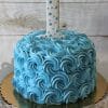 The CB-023G 6" Smash Cake features blue rosette icing and a white number one with blue polka dots on top, perfect for a first birthday celebration. It sits elegantly on a white cake stand.