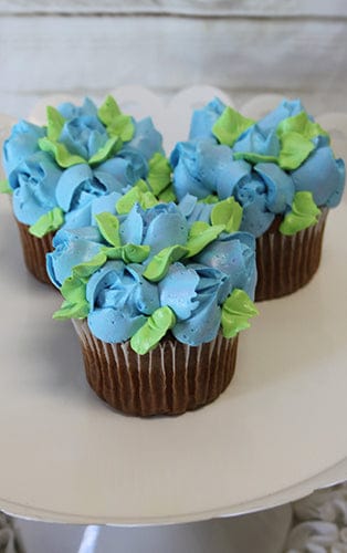 Three CC-090 Chocolate cupcakes with white filling, topped with blue and green petal-shaped icing to resemble hydrangea flowers, are arranged on a white plate for a floral presentation.