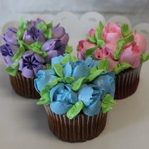 A CC-091 Gold cupcake with white filling is displayed on a white scalloped-edge plate, decorated with colorful buttercream flowers—purple, blue, and pink petals—and green leaves.