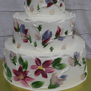 The WD-001W is a three-tiered white cake adorned with hand-painted red, purple, pink, and green floral designs, displayed on a gold cake board against a light wood background.