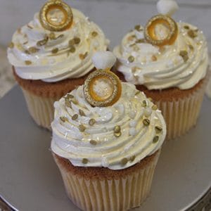 Three CC-094 Gold cupcakes with chocolate filling, featuring swirled white frosting, gold and pearl sprinkles, and fondant rings, displayed on a silver stand.