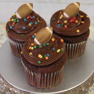 Three CC-095B chocolate cupcakes with white filling, swirled chocolate frosting, colorful round sprinkles, and small football-shaped decorations are displayed on a silver cake stand.