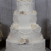 The WC-002A is a four-tier white wedding cake with intricate piping and white and pale pink roses on each tier, displayed against a rustic wooden backdrop.