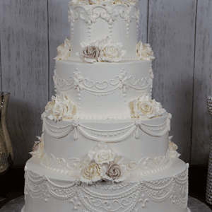 The WC-002A is a four-tier white wedding cake with intricate piping and white and pale pink roses on each tier, displayed against a rustic wooden backdrop.