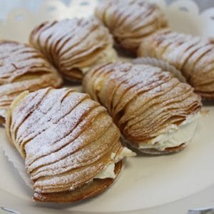 AP-046E Lobster Tail pastries, featuring a layered shell-like shape filled with cream and dusted with powdered sugar, are displayed on a white scalloped tray.