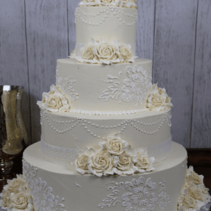The WC-060 is a four-tiered white wedding cake featuring piped lace designs, pearl-like dots, and clusters of white buttercream roses on each tier, beautifully displayed against a wooden backdrop.