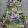 The WD-001X is a two-tiered pastel green wedding cake adorned with white, pink, and blue sugar flowers, green leaves, ornate white piping, and features a gold plaque on the top tier displaying the initials P & S.