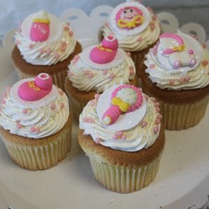 CC-111 Chocolate cupcakes with white filling, topped with swirled white icing, pink and white sugar pearls, and fondant baby-themed decorations like rattles and bottles, arranged on a white scalloped-edge plate.