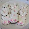 Six CE-040e Easter bunny-shaped cookies with white icing, pink noses, and pink paw details are arranged on a white scalloped-edge plate.