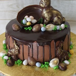 EA-027 is a chocolate cake with dark chocolate ganache drip, topped with a large chocolate bunny, an open egg filled with candy eggs, and decorated with smaller bunnies, candy eggs, and green icing leaves on a gold board.
