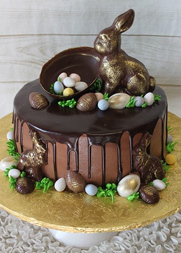EA-027 is a chocolate cake with dark chocolate ganache drip, topped with a large chocolate bunny, an open egg filled with candy eggs, and decorated with smaller bunnies, candy eggs, and green icing leaves on a gold board.