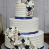 The WC-001A is a three-tiered white cake with textured icing, adorned with blue, white, and blush pink buttercream roses, green leaves, and finished with deep blue satin ribbons on each tier.