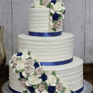 The WC-001A is a three-tiered white cake with textured icing, adorned with blue, white, and blush pink buttercream roses, green leaves, and finished with deep blue satin ribbons on each tier.