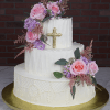 The RC-001D is a three-tiered white cake with textured icing, adorned with pink and purple flowers, greenery, and a gold cross on the middle tier, set on a gold cake board against a brick wall background.