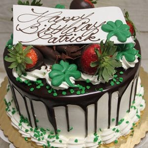A chocolate-drizzled birthday cake (Product: STP-005) with strawberries, green clover decor, whipped cream, and a "Happy Birthday Patrick" white chocolate plaque sits on a gold tray.