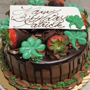 STP-006: Chocolate cake with ganache drip, chocolate-covered strawberries, green shamrock decorations, a white “Happy Birthday Patrick” plaque in brown script, and green sprinkles around the edges.