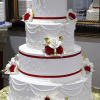 WC-061 is a three-tiered white wedding cake adorned with red and white roses, gold leaves, and red ribbons, presented on a gold cake board.