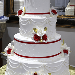 WC-061 is a three-tiered white wedding cake adorned with red and white roses, gold leaves, and red ribbons, presented on a gold cake board.