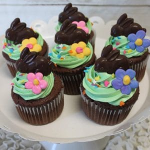 EA-042 Chocolate cupcakes with white filling, topped with green frosting, colorful flower decorations, and chocolate bunny figures, arranged on a white cake stand.