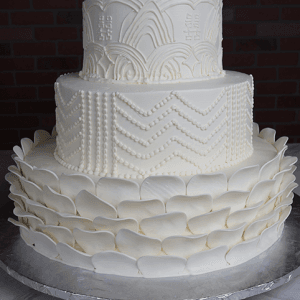 The WD-001Y is a three-tiered white cake featuring intricate scalloped icing designs, piped dots, and petal-like decorations at the base, set against a dark brick background.