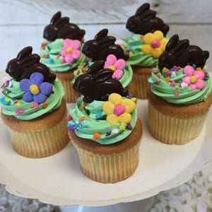 Six EA-043 Gold cupcakes with chocolate filling, topped with green frosting, flower decorations, sprinkles, and chocolate bunny figures are arranged on a white cake stand against a light wood background.