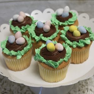 Six EA-045 Gold cupcakes with chocolate filling, topped with chocolate frosting, green icing like grass, and candy eggs in a bird nest style, are displayed on a white scalloped cake stand.