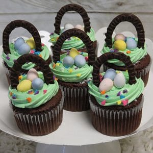 Five EA-047 Gold cupcakes with chocolate filling, decorated with green frosting, chocolate handles, pastel candy eggs, and colorful heart-shaped sprinkles to resemble Easter baskets, displayed on a white cake stand.