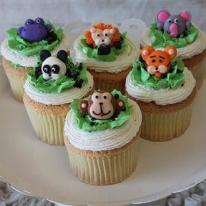 Six CC-104 gold cupcakes with chocolate filling are topped with fondant animals—hippo, lion, elephant, panda, tiger, and monkey—and displayed on a white cake stand.