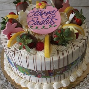 A Gold Cake with Lemon and Raspberry mousse filling, featuring striped sides, topped with strawberries, chocolate-dipped strawberries, lemon slices, white chocolate curls, and a pink fondant disc that says "Happy Easter" in purple icing.