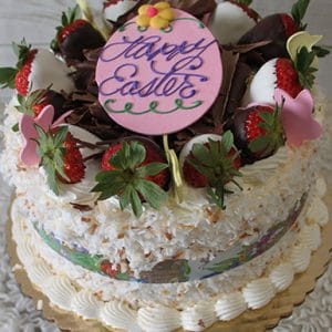 The Pina Colada Cake is a festive treat with chocolate-dipped strawberries, chocolate shavings, pastel flower decorations, a pink Happy Easter plaque, coconut-covered sides, and a piped cream border.