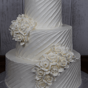 The WC-001B is a three-tiered white wedding cake with textured diagonal lines, adorned with clusters of white buttercream roses. Displayed on a silver board, it stands out beautifully against a wooden background.