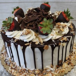 The BD-018A Almond Joy Cake is a round cake with white frosting, chocolate ganache drips, topped with chocolate-dipped strawberries, whipped cream swirls, chocolate shavings, and toasted almonds and coconut around the base.