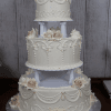 The WC-002B is a three-tiered white wedding cake featuring intricate icing, cream and pale pink rose decorations, showcased on a wooden table against a rustic wood panel backdrop.