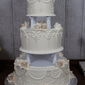 The WC-002B is a three-tiered white wedding cake featuring intricate icing, cream and pale pink rose decorations, showcased on a wooden table against a rustic wood panel backdrop.