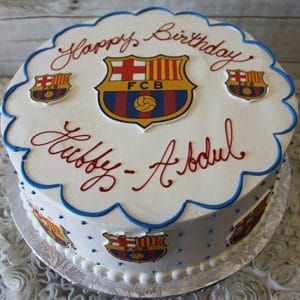 The ST-007A Futbol Club Barcelona round white cake features FC Barcelona logos, blue piping, and "Happy Birthday Hubby - Abdul" in red on top. It sits on a silver board over a floral tablecloth.