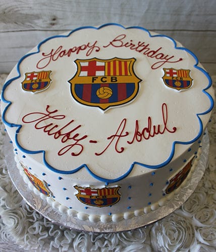 The ST-007A Futbol Club Barcelona round white cake features FC Barcelona logos, blue piping, and "Happy Birthday Hubby - Abdul" in red on top. It sits on a silver board over a floral tablecloth.