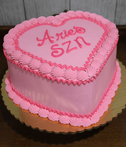 A BD-023A heart-shaped pink cake with piped frosting edges and "Aries SZN" in pink icing on top, presented on a gold cake board.