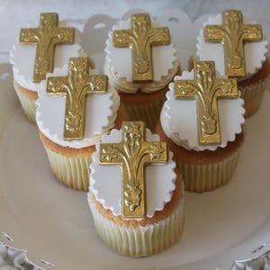 Six CC-043 Gold cupcakes with chocolate filling, decorated with white fondant and detailed gold crosses, arranged on a white scalloped-edge cake stand.