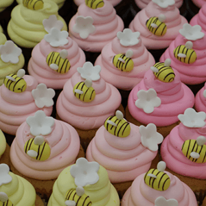 Rows of CC-140 Gold cupcakes with chocolate filling feature pink or yellow frosting, fondant bees, and white flower decorations. The cupcakes are arranged closely together in a box.